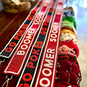 May include: Red and black beaded straps with the text "GO POKES BOOMER" and "GO POKES BOOMER SOONER" repeated. The straps are on a wooden surface.