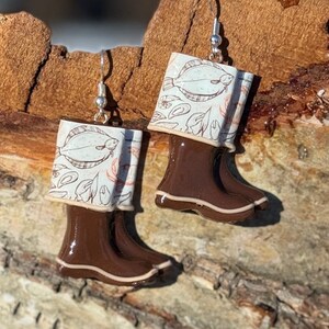 May include: A pair of earrings featuring brown rain boots with a cream-colored cuff. The cuff has a pattern of fish and leaves in shades of cream and orange. The earrings hang from silver-colored hooks.