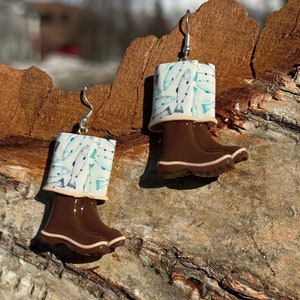 May include: A pair of earrings shaped like brown wellington boots with a white top. The white top has a blue fish pattern. The earrings are suspended from silver hooks.
