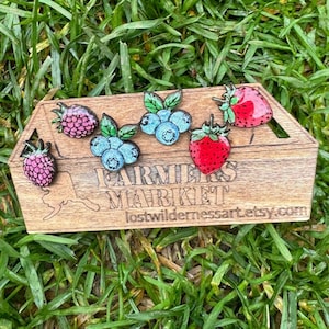 May include: Wooden box with berry-shaped pins. The pins include raspberries, blueberries, and strawberries. The box has the words "FARMERS MARKET" and the Etsy shop URL "lostwildernessart.etsy.com" engraved on it.
