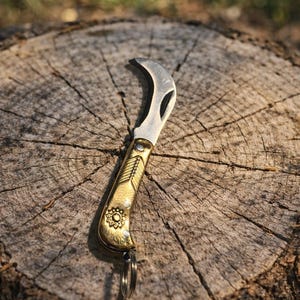 Puede incluir: Una pequeña navaja de podar curvada con una hoja plateada y un mango dorado. El mango presenta un diseño floral en relieve. Un llavero está unido a la base del mango. La navaja descansa sobre un tocón de árbol desgastado.