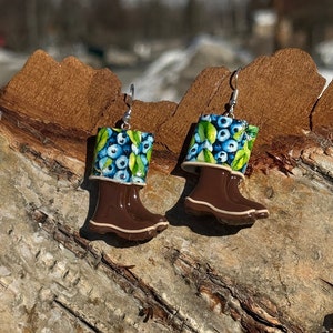 May include: A pair of whimsical earrings shaped like brown rain boots. The top of each boot features a pattern of blue blueberries and green leaves. The earrings hang from silver-colored hooks.