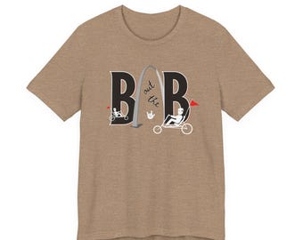 May include: A heathered tan short-sleeve t-shirt featuring a graphic with two large black letters B, a gray arch, and the text "out the" with a hand sign. Two white stick figures ride recumbent trikes with small red flags on the sides.
