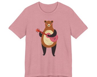 May include: A heathered mauve short-sleeve t-shirt featuring a centered graphic of a brown bear with a cream-colored belly, standing upright and playing a red acoustic guitar. The bear has black limbs and a simple, friendly facial expression.