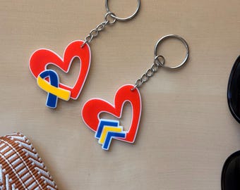May include: Two red heart-shaped keychains with a blue and yellow ribbon design. The keychains are connected to silver key rings with a chain. The keychains are on a light brown surface.