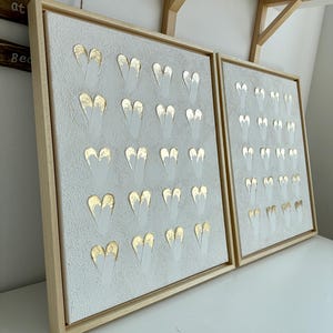May include: Two framed canvas artworks. Each has a textured, light grey background with a pattern of white and gold heart shapes. The frames are light-coloured wood, and the art is displayed on a white surface.