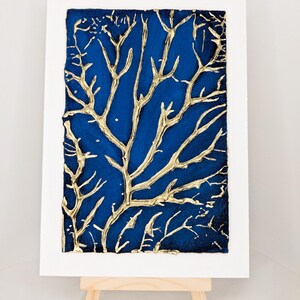May include: A gold leaf tree design on a deep blue background. The artwork is framed and displayed on a small wooden easel.