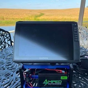 K&ouml;nnte beinhalten: Ein schwarzer Garmin Fischfinder mit einem blauen und schwarzen St&auml;nder. Der St&auml;nder hat einen Spannungswandler und eine Batterie. Der Fischfinder steht auf einem Tisch mit einer blauen Tischdecke. Der Hintergrund ist ein Maisfeld.