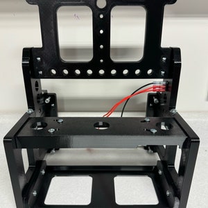 K&ouml;nnte beinhalten: Schwarzer Metallrahmen mit einer rechteckigen &Ouml;ffnung und einer kleineren rechteckigen &Ouml;ffnung oben. Der Rahmen hat mehrere L&ouml;cher und ist f&uuml;r die Aufnahme eines Ger&auml;ts ausgelegt.