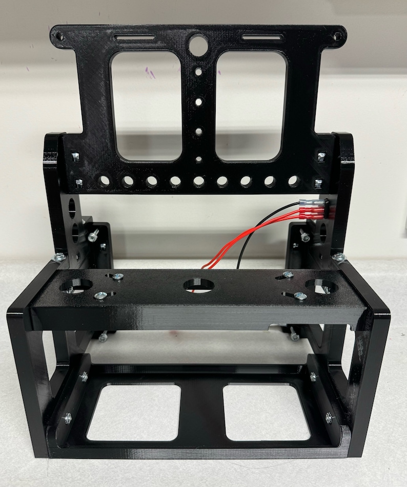 K&ouml;nnte beinhalten: Schwarzer Metallrahmen mit einer rechteckigen &Ouml;ffnung und einer kleineren rechteckigen &Ouml;ffnung oben. Der Rahmen hat mehrere L&ouml;cher und ist f&uuml;r die Aufnahme eines Ger&auml;ts ausgelegt.