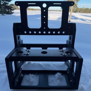 May include: A black metal bracket with a rectangular base and an open design. The bracket has multiple holes and slots, and is secured with silver screws. The background shows a snowy landscape and a tree.
