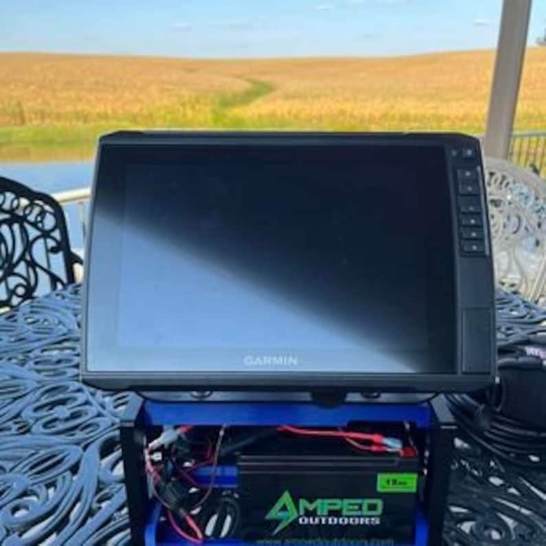 K&ouml;nnte beinhalten: Ein schwarzer Garmin Fischfinder mit einem blauen und schwarzen St&auml;nder. Der St&auml;nder hat einen Spannungswandler und eine Batterie. Der Fischfinder steht auf einem Tisch mit einer blauen Tischdecke. Der Hintergrund ist ein Maisfeld.