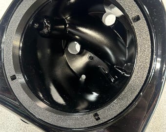 May include: A close-up shot of a black, glossy, angular object with a circular opening. The opening has a gray, textured ring around it. The interior is black with several circular holes and a central, cylindrical component.
