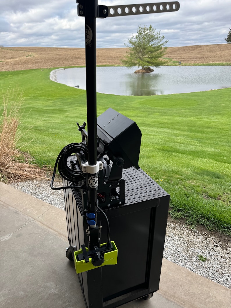 K&ouml;nnte beinhalten: Ein schwarzer Metallwagen mit einer schwarzen Leuchte, die an einem Mast befestigt ist. Der Wagen hat eine schwarze Metalloberseite mit einem Diamantplattenmuster. Die Leuchte hat ein schwarzes Metallgeh&auml;use und einen wei&szlig;en Kunststoffgriff. Der Wagen steht auf einer Betonfl&auml;che vor einer gr&uuml;nen Grasfl&auml;che mit einem Teich im Hintergrund.