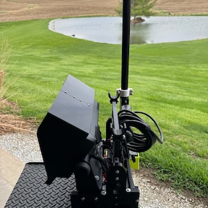 K&ouml;nnte beinhalten: Ein schwarzer Monitor mit einem Sonnenschutz, der auf einem schwarzen Metallst&auml;nder montiert ist. Der Monitor ist mit einem schwarzen Kabel verbunden und vor einem gr&uuml;nen Grasfeld mit einem Teich im Hintergrund positioniert.