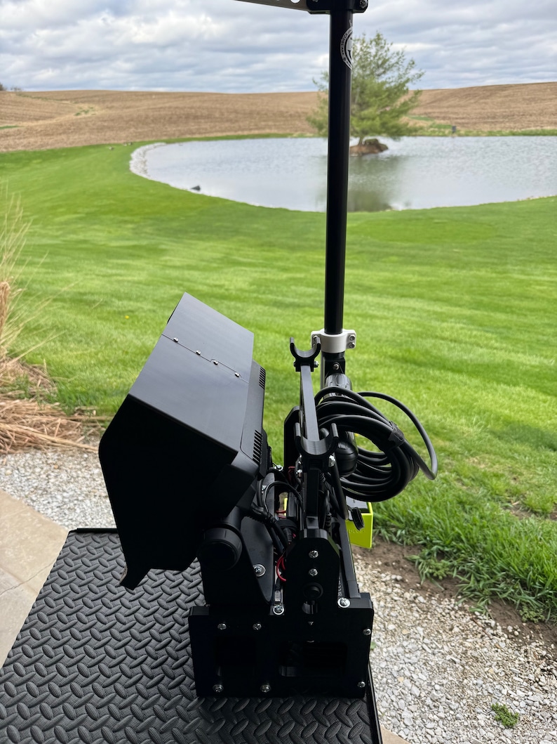 K&ouml;nnte beinhalten: Ein schwarzer Monitor mit einem Sonnenschutz, der auf einem schwarzen Metallst&auml;nder montiert ist. Der Monitor ist mit einem schwarzen Kabel verbunden und vor einem gr&uuml;nen Grasfeld mit einem Teich im Hintergrund positioniert.