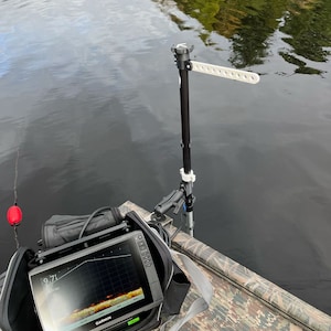 May include: A black and gray fish finder with a screen displaying a sonar reading. The fish finder is in a black carrying case and mounted on a black pole with a white strap. The pole is mounted on a boat.