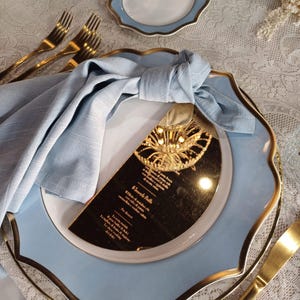 May include: A blue and gold scalloped plate with a gold rim. A blue linen napkin is folded on the plate. A gold mirror with a menu printed on it is on top of the napkin. The menu is in Italian and includes the text "Il Secondo Piatto".