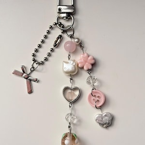 May include: A silver-toned keychain with a lobster clasp, featuring a cross charm, a chain, and various decorative elements. These include a pink flower, a heart-shaped charm, a button, and a strawberry-shaped bead. The keychain also has a Hello Kitty charm.