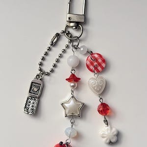 May include: A silver keychain with a lobster clasp and a dangling charm arrangement. The charms include a miniature flip phone, white and red beads, a star, a heart, a flower, and a red apple with a yellow heart cutout.
