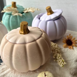 Könnte beinhalten: Drei Keramikbehälter in Kürbisform mit Deckeln. Die Kürbisse sind in Pastellfarben: hellblau, Lavendel und hellrosa. Die Deckel haben goldene Akzente.