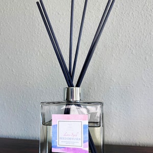 May include: A clear glass bottle with a silver lid and black reeds. The bottle has a label that says "Love Spell Reed Diffuser" with a watercolor design of pink, blue, and purple.