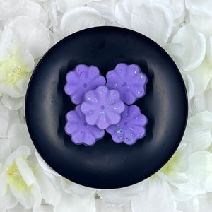 May include: Five purple wax melts shaped like flowers with a glittery finish, displayed on a black plate.