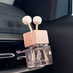 Könnte beinhalten: Ein hölzerner Autolufterfrischer mit zwei weißen Diffusorstäbchen. Die Flasche ist klar und mit einer Flüssigkeit gefüllt. Der Lufterfrischer ist an einem Lüftungsschlitz im Auto befestigt.