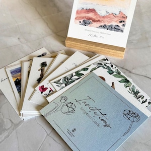 May include: A set of scripture cards with watercolor illustrations. The cards feature various designs, including floral patterns and landscape paintings. The text "Invitations Scripture Cards" is visible on the front of a card. The phrase "Let them hear" is written on the top of a card.