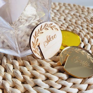 Puede incluir: Dos porta tarjetas de lugar redondos, uno de madera con un dise&ntilde;o de hojas y el nombre "Steve" grabado, el otro de acr&iacute;lico espejo dorado con el nombre "Emily" grabado.
