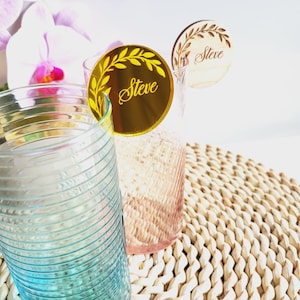 Puede incluir: Dos marcadores de bebida de acr&iacute;lico dorados con un dise&ntilde;o de hojas y el nombre "Steve" escrito en letras doradas. Los marcadores se colocan encima de dos vasos transparentes con un dise&ntilde;o texturizado.