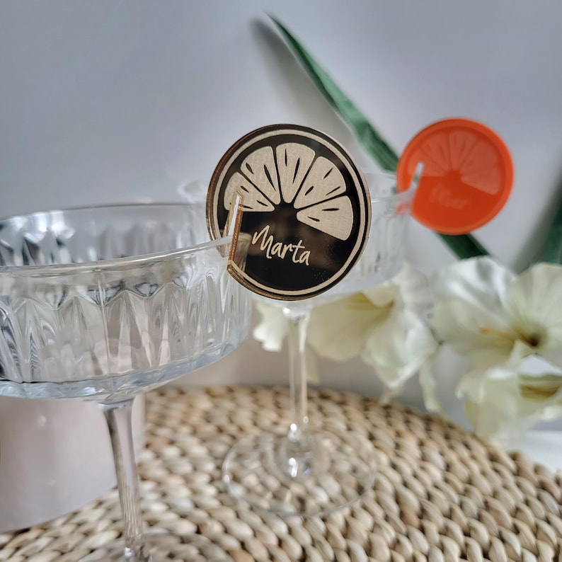 May include: Two clear glass champagne flutes with wooden drink markers. The markers are shaped like a lemon slice and have the name "Marta" written on them.