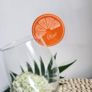 Puede incluir: Marcador de bebida de acrílico en forma de naranja con el nombre "Oliver" grabado en él. El marcador se coloca en el borde de un vaso transparente.