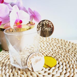 Puede incluir: Dos porta tarjetas de lugar dorados con un dise&ntilde;o de hojas. Las tarjetas de lugar est&aacute;n grabadas con los nombres "Emily" y "Steve".