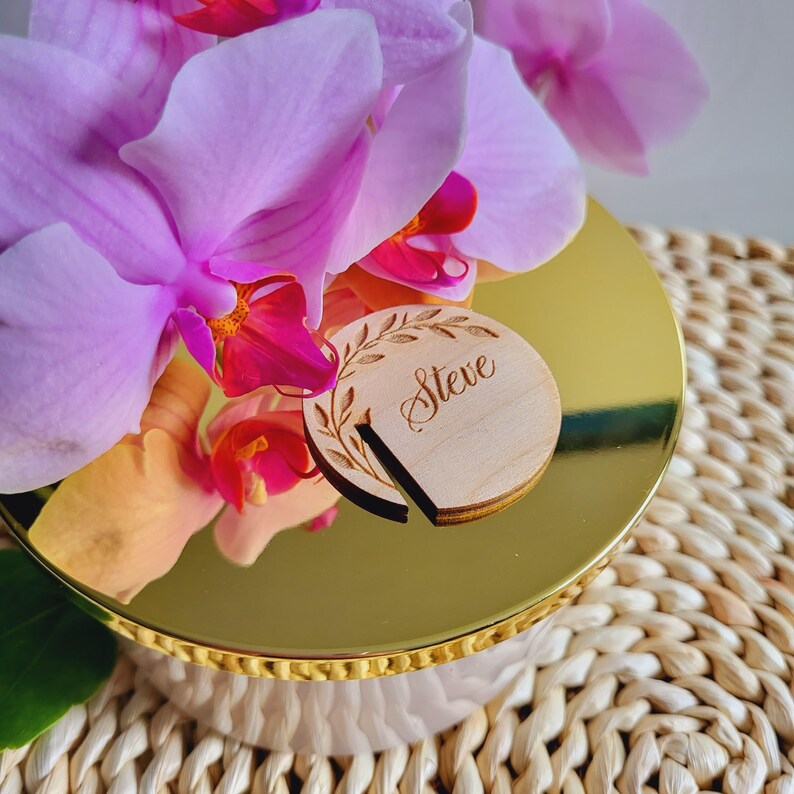 Puede incluir: Un tarro de vela dorado con tapa y una etiqueta de nombre de madera que dice "Steve" con un dise&ntilde;o floral. El tarro de vela est&aacute; rodeado de flores de orqu&iacute;dea rosa.