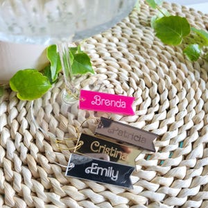 May include: Four acrylic wine glass charms with names printed on them. The charms are in the shape of a banner. The names are Brenda, Patricia, Cristina, and Emily. The charms are pink, silver, gold, and black.