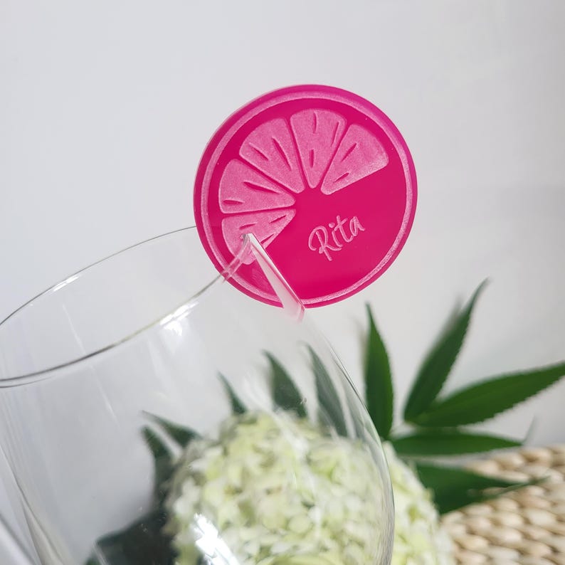 May include: Pink acrylic drink marker with a lemon slice design and the name "Rita" written on it. The marker is placed on a clear wine glass.