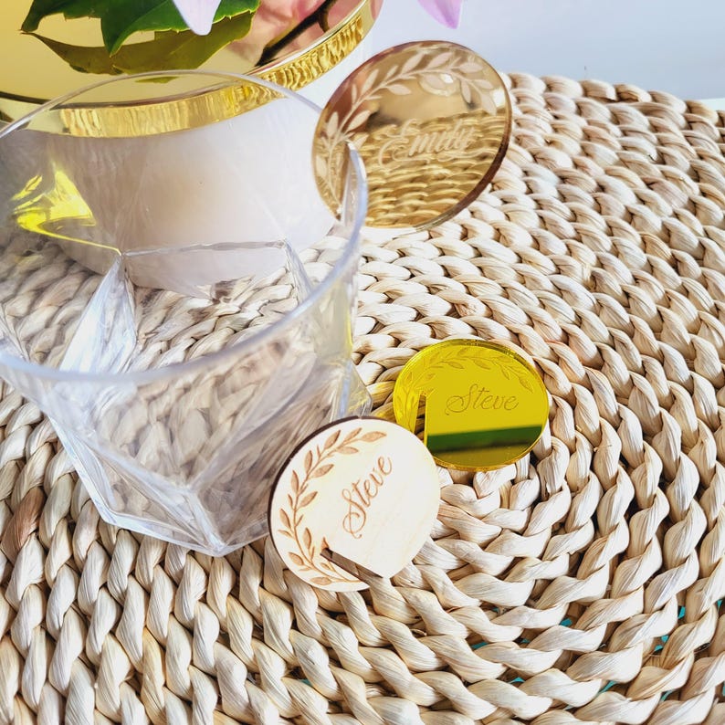 Puede incluir: Dos porta tarjetas de lugar dorados con los nombres "Emily" y "Steve" grabados en ellos. Un porta tarjetas de lugar es de madera con un dise&ntilde;o de hojas y el otro es de vidrio espejo.