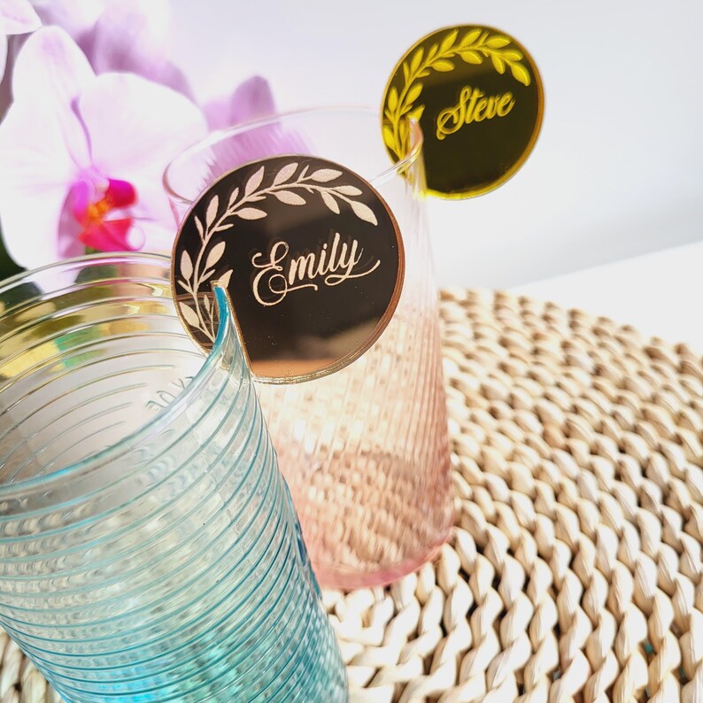 Puede incluir: Dos marcadores de bebida de acr&iacute;lico espejo dorado con un dise&ntilde;o de hojas y los nombres "Emily" y "Steve" escritos en cursiva.