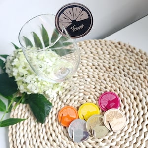 May include: A set of six lemon-shaped drink markers with names written on them. The markers are made of wood and have a gold finish. The markers are sitting on a woven placemat.