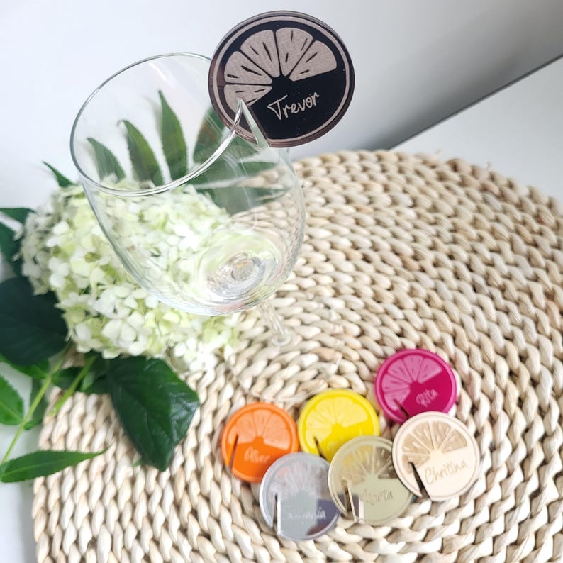 May include: A set of six lemon-shaped drink markers with names written on them. The markers are made of wood and have a gold finish. The markers are sitting on a woven placemat.