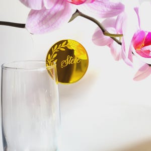 Puede incluir: Tarjeta de lugar de espejo dorado con un dise&ntilde;o de hojas y el nombre "Steve" grabado en ella. La tarjeta de lugar est&aacute; sobre una superficie blanca.