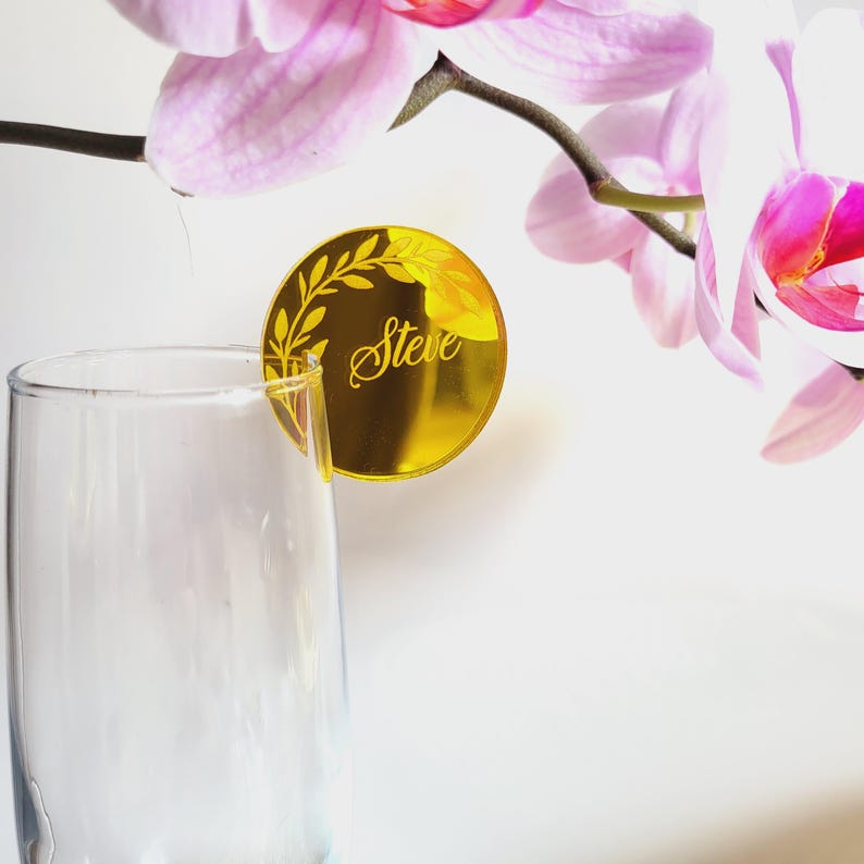 Puede incluir: Tarjeta de lugar de espejo dorado con un dise&ntilde;o de hojas y el nombre "Steve" grabado en ella. La tarjeta de lugar est&aacute; sobre una superficie blanca.