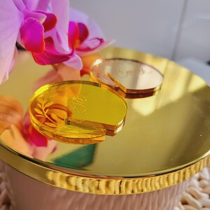 Puede incluir: Dos tapas de tapa de vela de acr&iacute;lico de color dorado con texto grabado. La tapa de la izquierda dice "Ella" y tiene un dise&ntilde;o de hoja. La tapa de la derecha dice "Emily".