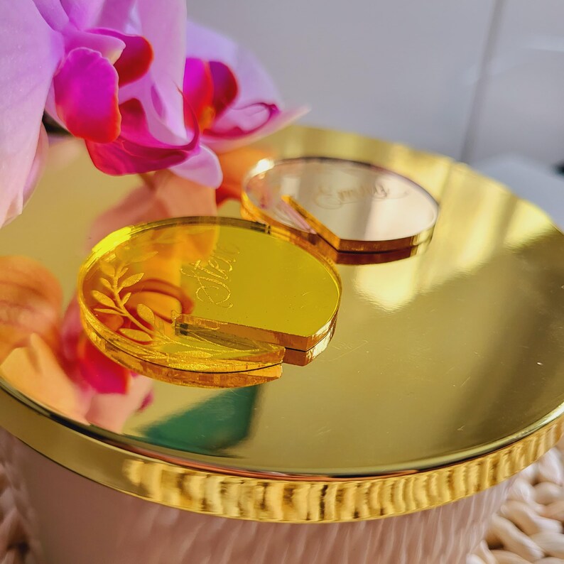 Puede incluir: Dos tapas de tapa de vela de acr&iacute;lico de color dorado con texto grabado. La tapa de la izquierda dice "Ella" y tiene un dise&ntilde;o de hoja. La tapa de la derecha dice "Emily".
