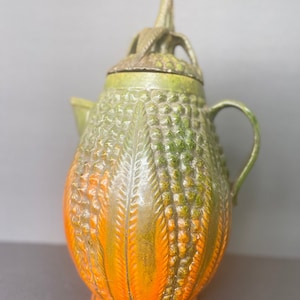 Könnte beinhalten: Eine grüne und orangefarbene Teekanne aus Keramik mit einer strukturierten Oberfläche und einem dekorativen Deckel. Die Teekanne hat einen runden Körper und einen Ausguss auf einer Seite.