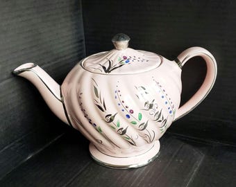 Può includere: Una teiera rosa chiaro con una texture a coste a spirale, dotata di bordo, manico, beccuccio e pomello del coperchio color argento. Il corpo è decorato con motivi a foglie nere dipinte e piccoli punti blu e viola in motivi verticali.