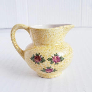 May include: A small, yellow ceramic pitcher with a speckled finish and a curved handle. The pitcher is decorated with three floral designs featuring pink and red roses with green leaves. The interior is white.