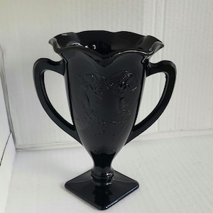 May include: A black glass vase with two handles and a ruffled rim. The vase features an embossed design of two figures and decorative elements. The vase sits on a square base and is a decorative item.