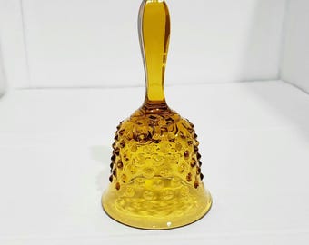 Peut inclure: Cloche en verre ambré avec une longue poignée incurvée. La cloche a une surface texturée avec de petits points en relief. L'objet est décoratif, probablement pour l'exposition ou la collection.